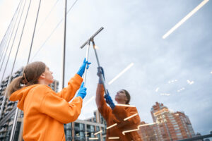 Young cleaning lady in a yellow hoodie and blue gloves cleans large windows with a long-handled window cleaning mop