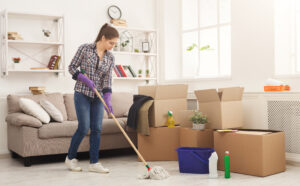 New home cleaning. Young woman tiding up after moving to new appartment. Girl with various detergents, rags and mops in living room full of carton boxes. Household and spring-cleanig concept